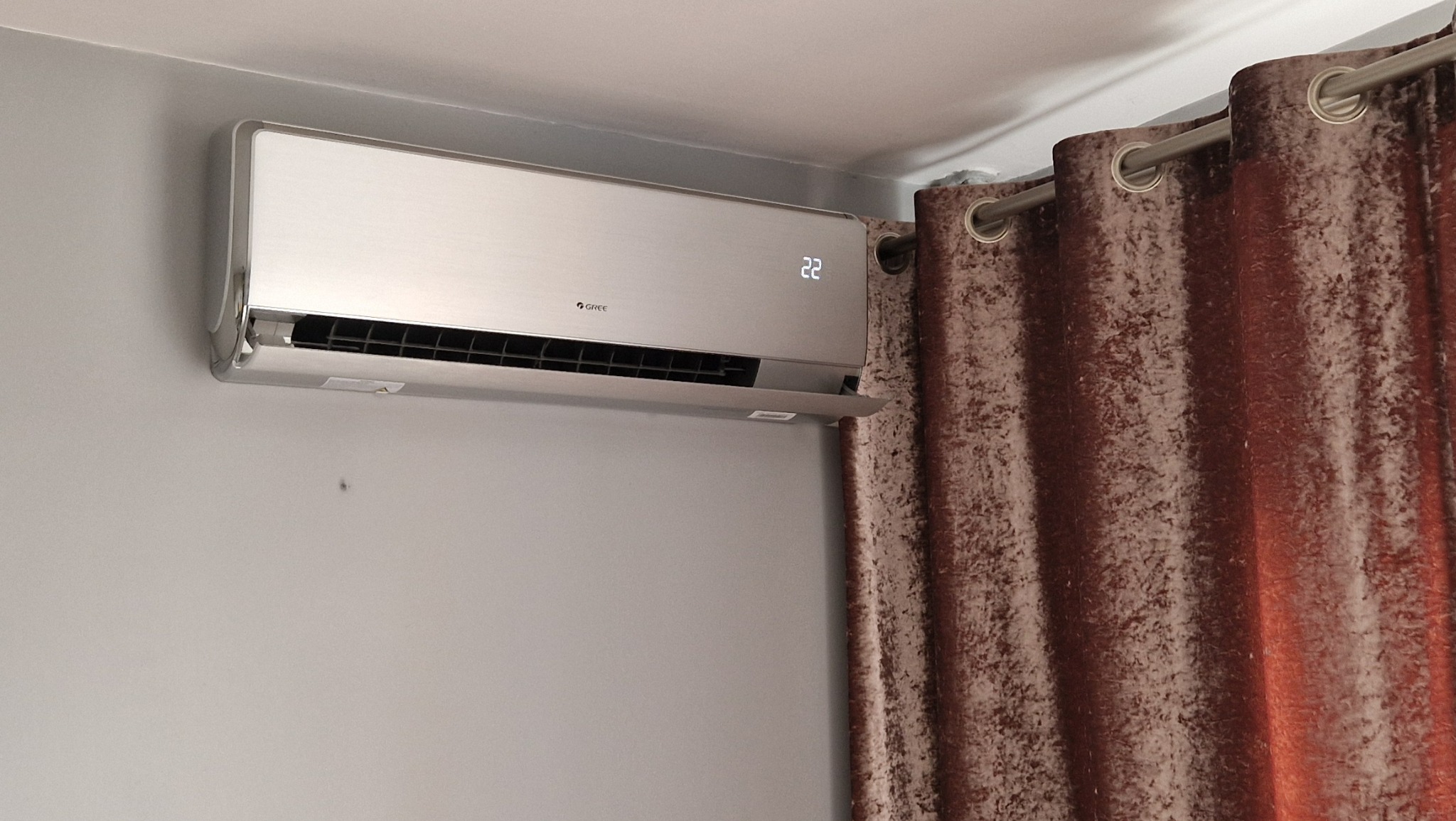 Srebrny klimatyzator Gree zamontowany na szarej ścianie, obok brązowych zasłon. Widoczny wyświetlacz z temperaturą 22 stopnie.
