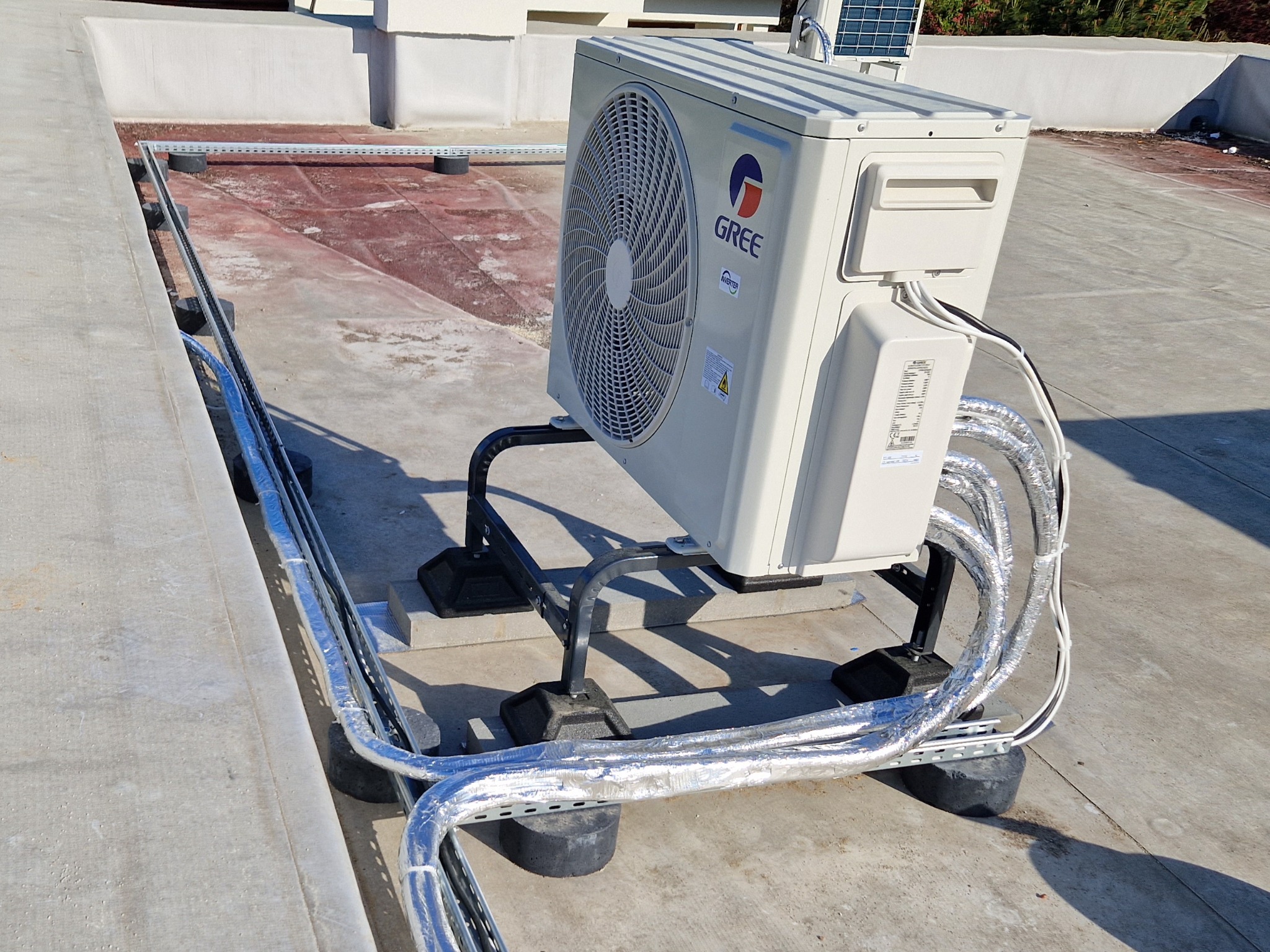 Biała jednostka zewnętrzna klimatyzacji marki Gree zamontowana na dachu, z widocznymi rurami w srebrnej izolacji i elementami mocującymi. Słoneczny dzień.