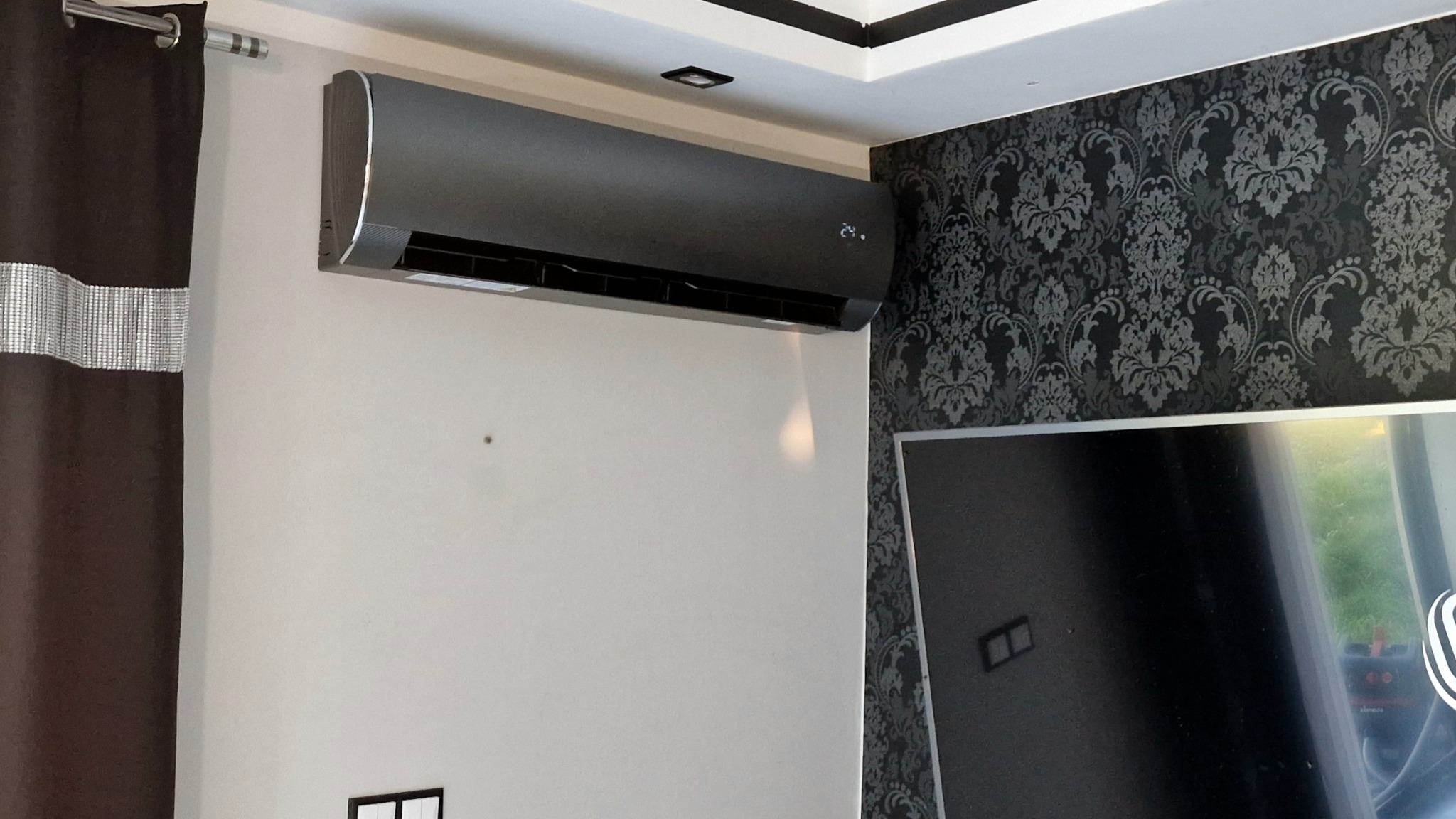 Nowoczesny, grafitowy klimatyzator ścienny zamontowany w pomieszczeniu z wzorzystą tapetą i ciemną zasłoną. Widoczny wyświetlacz temperatury.