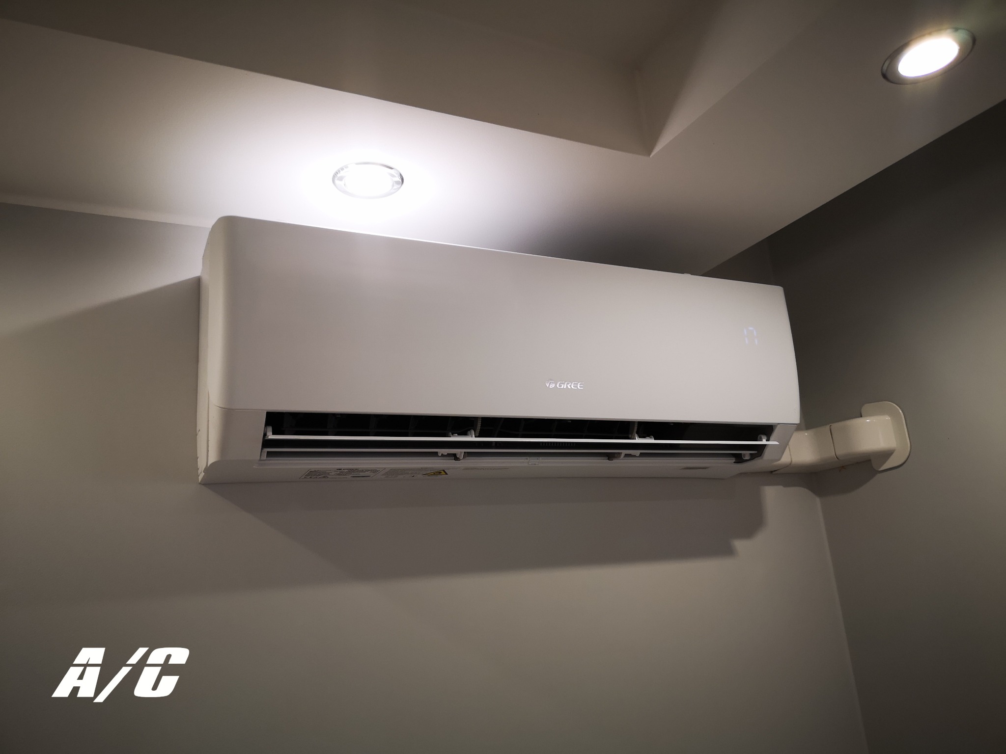 Biały klimatyzator GREE na szarej ścianie, widoczne przewody instalacyjne i oświetlenie sufitowe. W lewym dolnym rogu napis A/C.