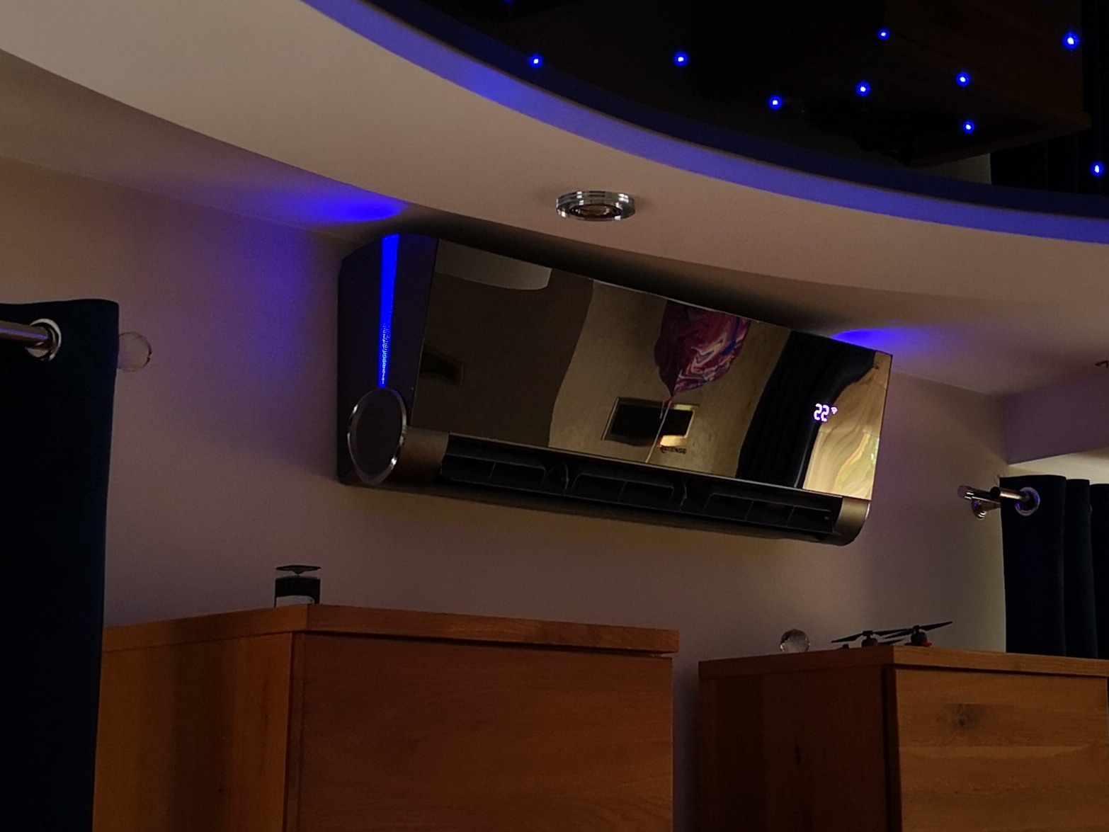 Nowoczesna, czarna klimatyzacja ścienna z niebieskim podświetleniem LED, zamontowana w pomieszczeniu z drewnianymi meblami i dekoracyjnym oświetleniem sufitowym.