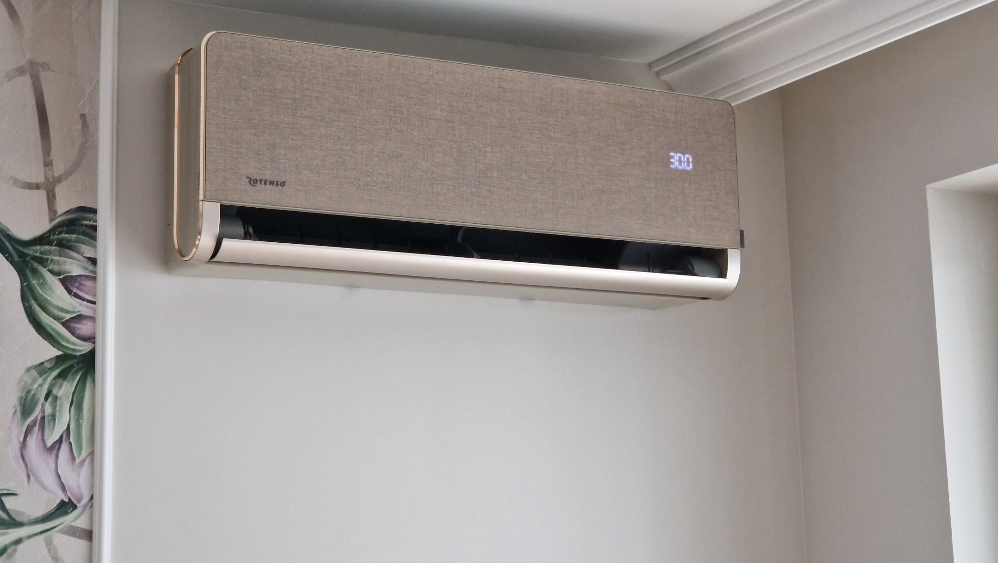 Beżowy klimatyzator Rotenso z wyświetlaczem 30.0 zamontowany na ścianie w jasnym wnętrzu, widoczny fragment dekoracyjnej tapety z motywem kwiatowym.