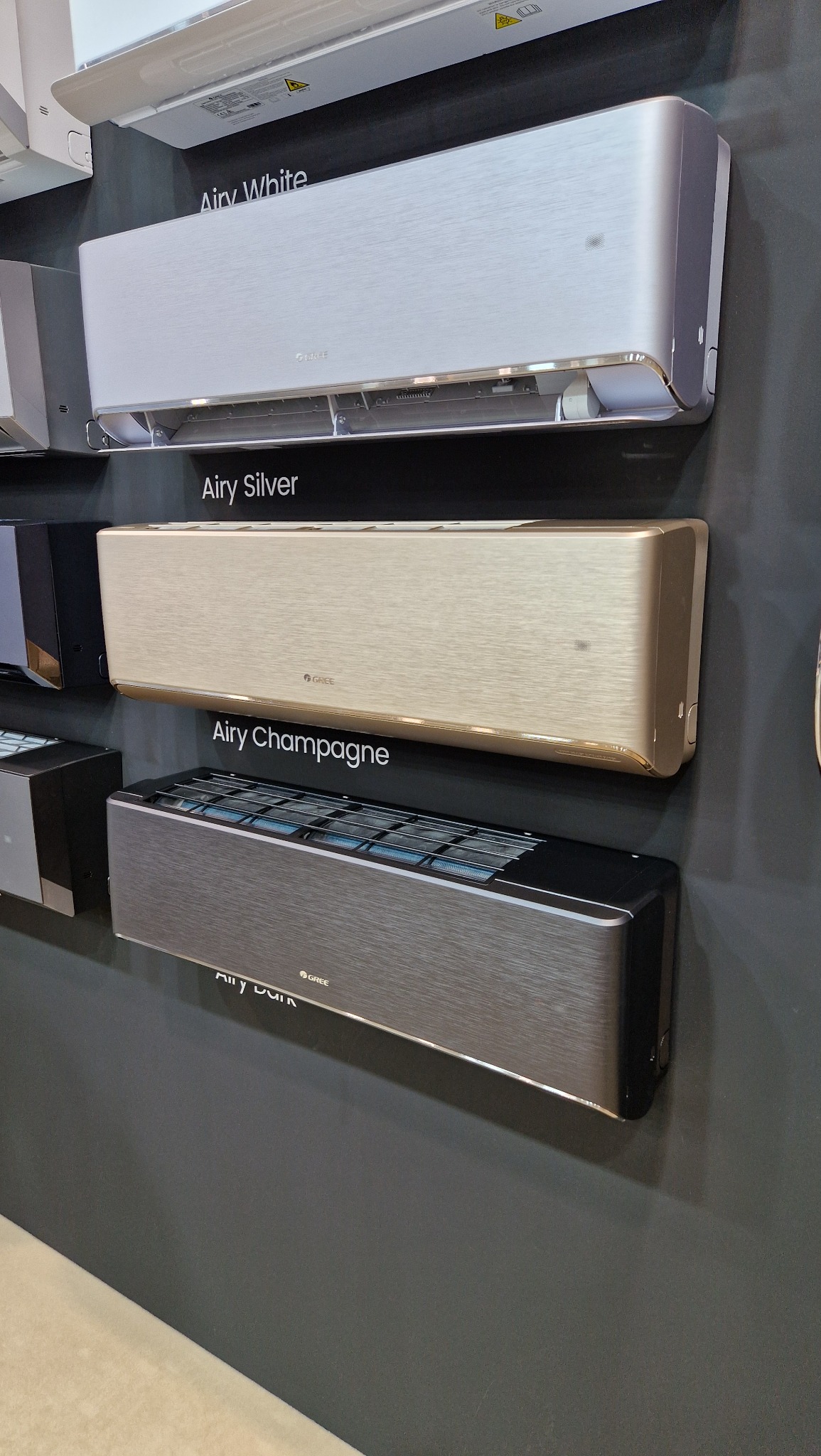Ekspozycja trzech modeli klimatyzatorów Gree Airy w odcieniach: White, Silver i Champagne, zamontowanych na ciemnoszarym tle. Widoczne detale wykończenia i logo producenta.