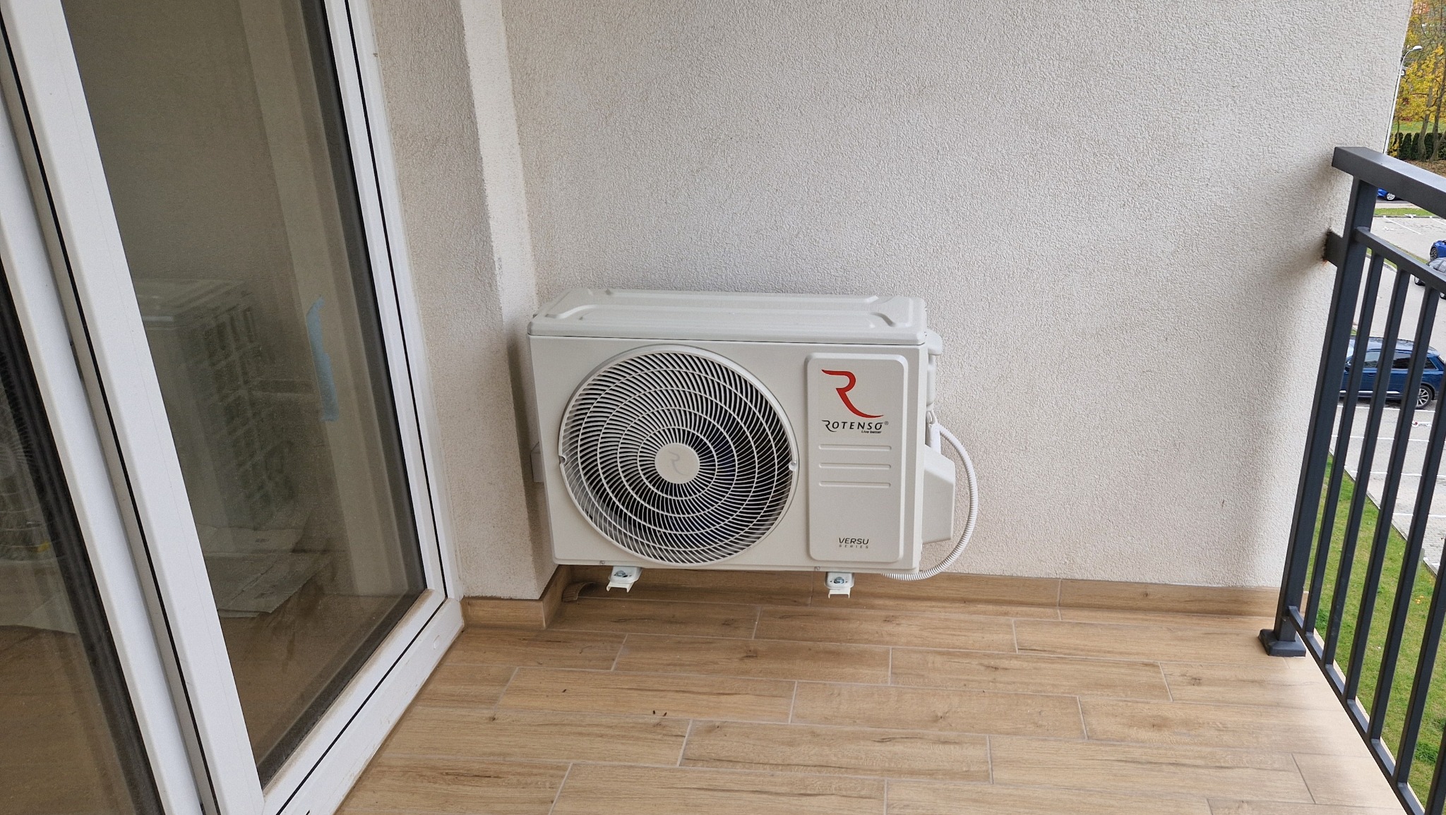 Biała jednostka zewnętrzna klimatyzacji Rotenso zamontowana na balkonie z terakotą, widok z boku, z fragmentem okna i balustrady w tle.