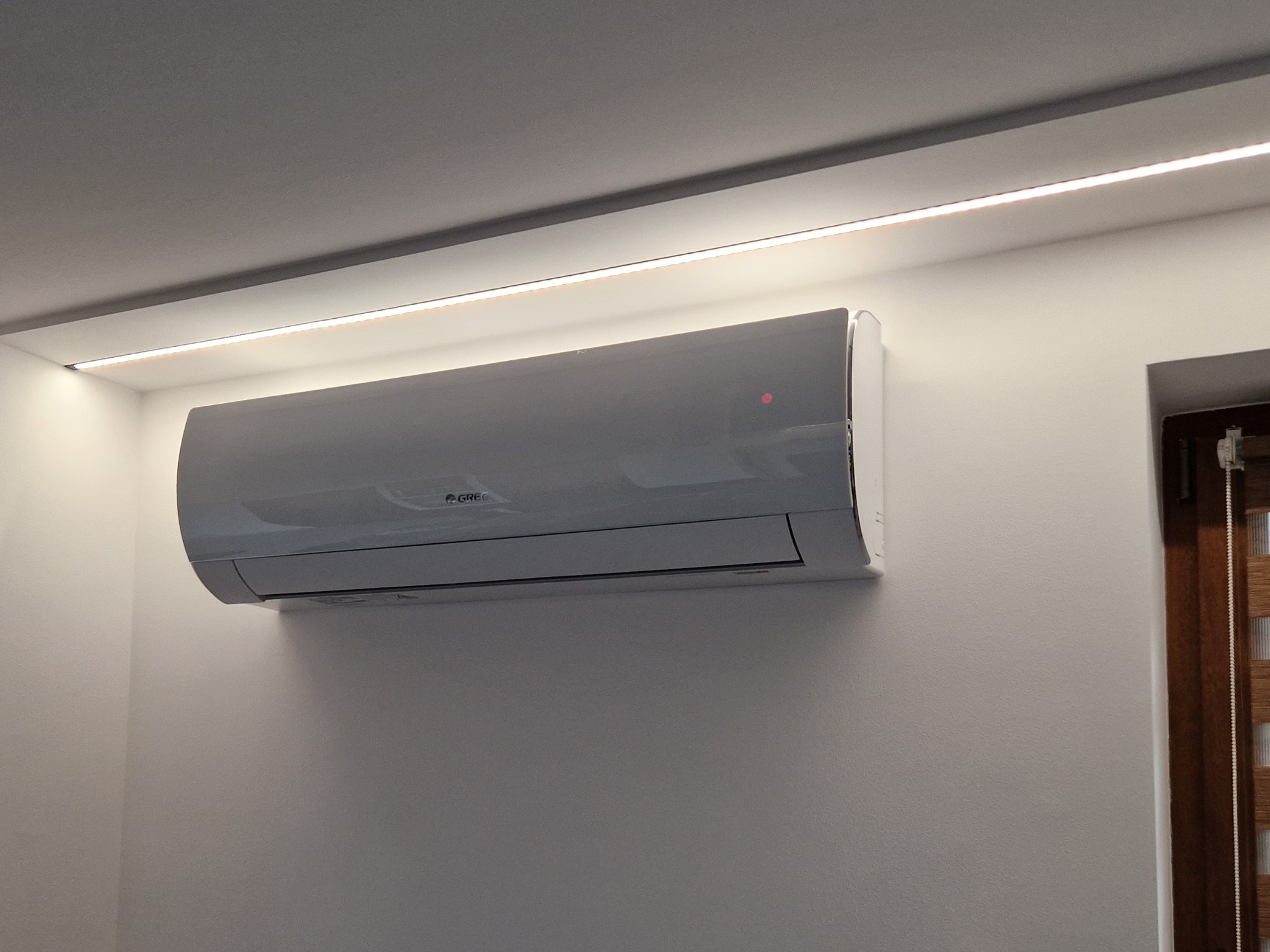 Zamontowany klimatyzator Gree na białej ścianie, oświetlony listwą LED przy suficie, z fragmentem okna z żaluzjami w tle. Nowoczesny design i minimalistyczny styl.