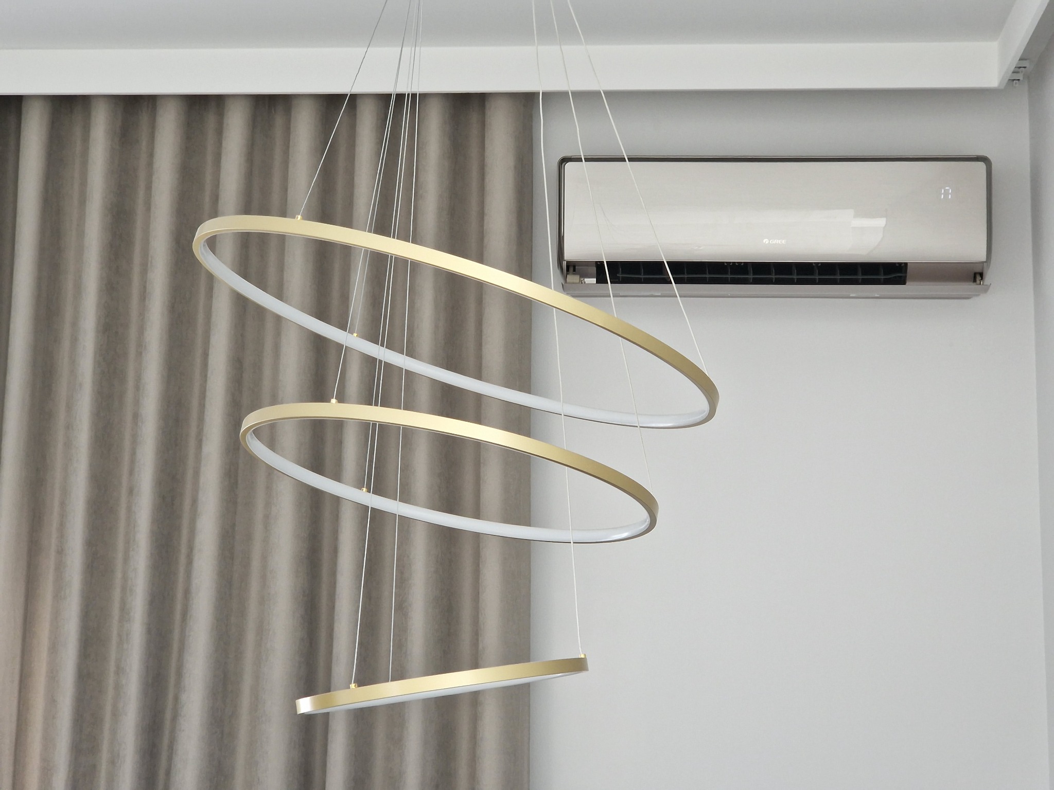 Zamontowany klimatyzator marki Gree na białej ścianie, obok zwisająca, nowoczesna lampa z trzema złotymi okręgami i szarą zasłoną w tle.