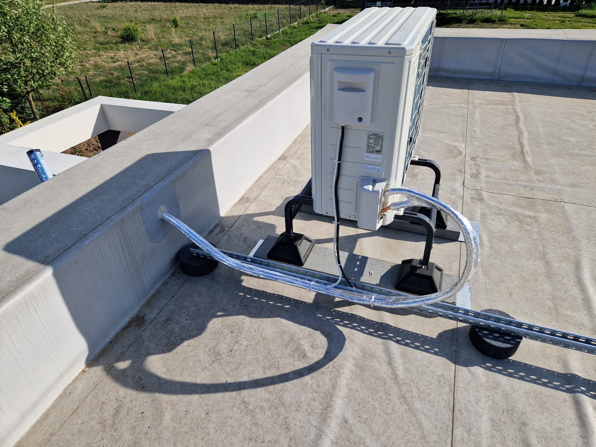 Zewnętrzna jednostka klimatyzacji zamontowana na płaskim dachu budynku, z widocznymi przewodami w osłonie i elementami montażowymi.