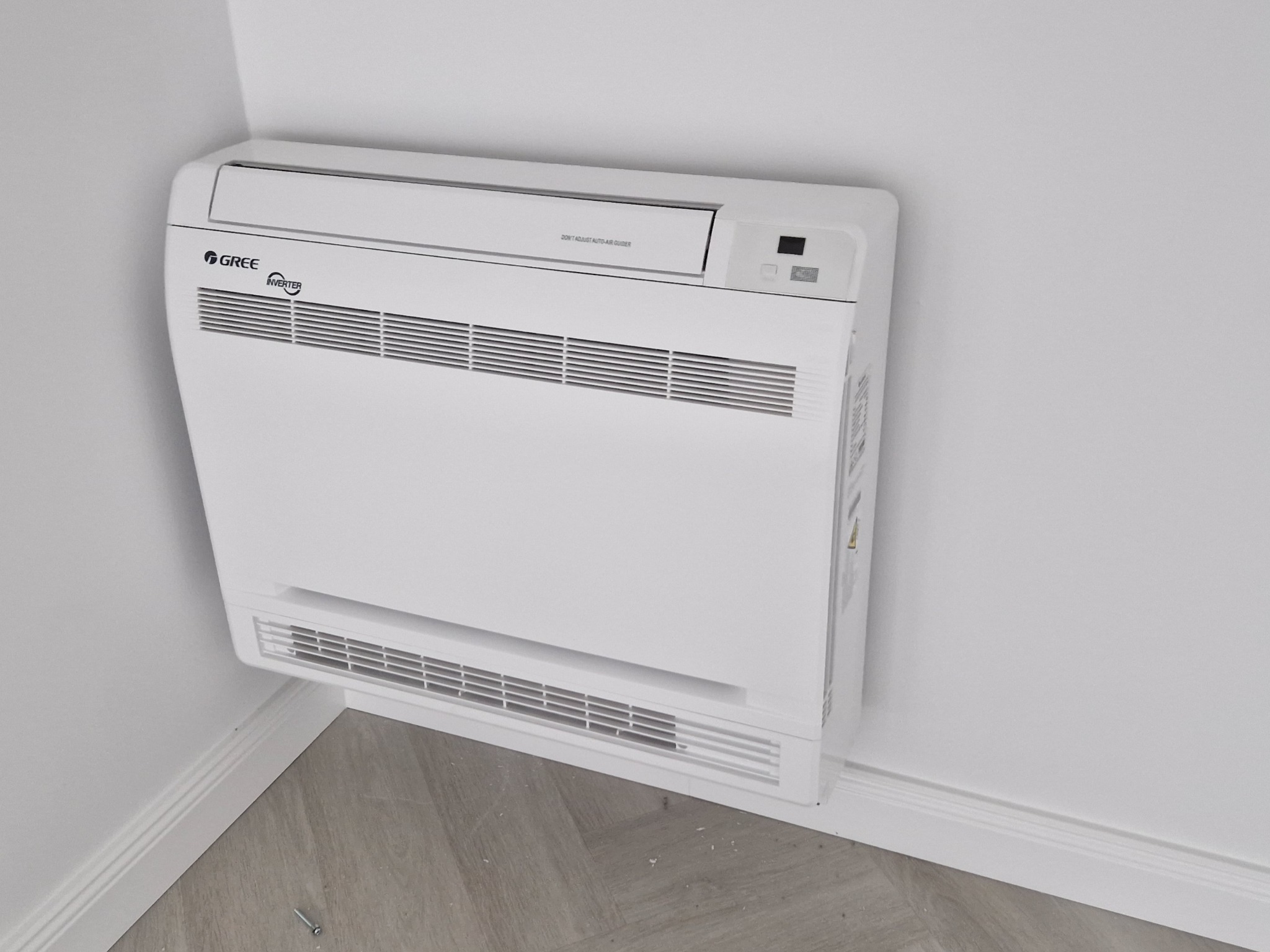 Biała, przypodłogowa jednostka wewnętrzna klimatyzacji marki Gree Inverter zamontowana w rogu pomieszczenia z szarymi panelami podłogowymi i białą listwą przypodłogową. Na podłodze leży śrubka.