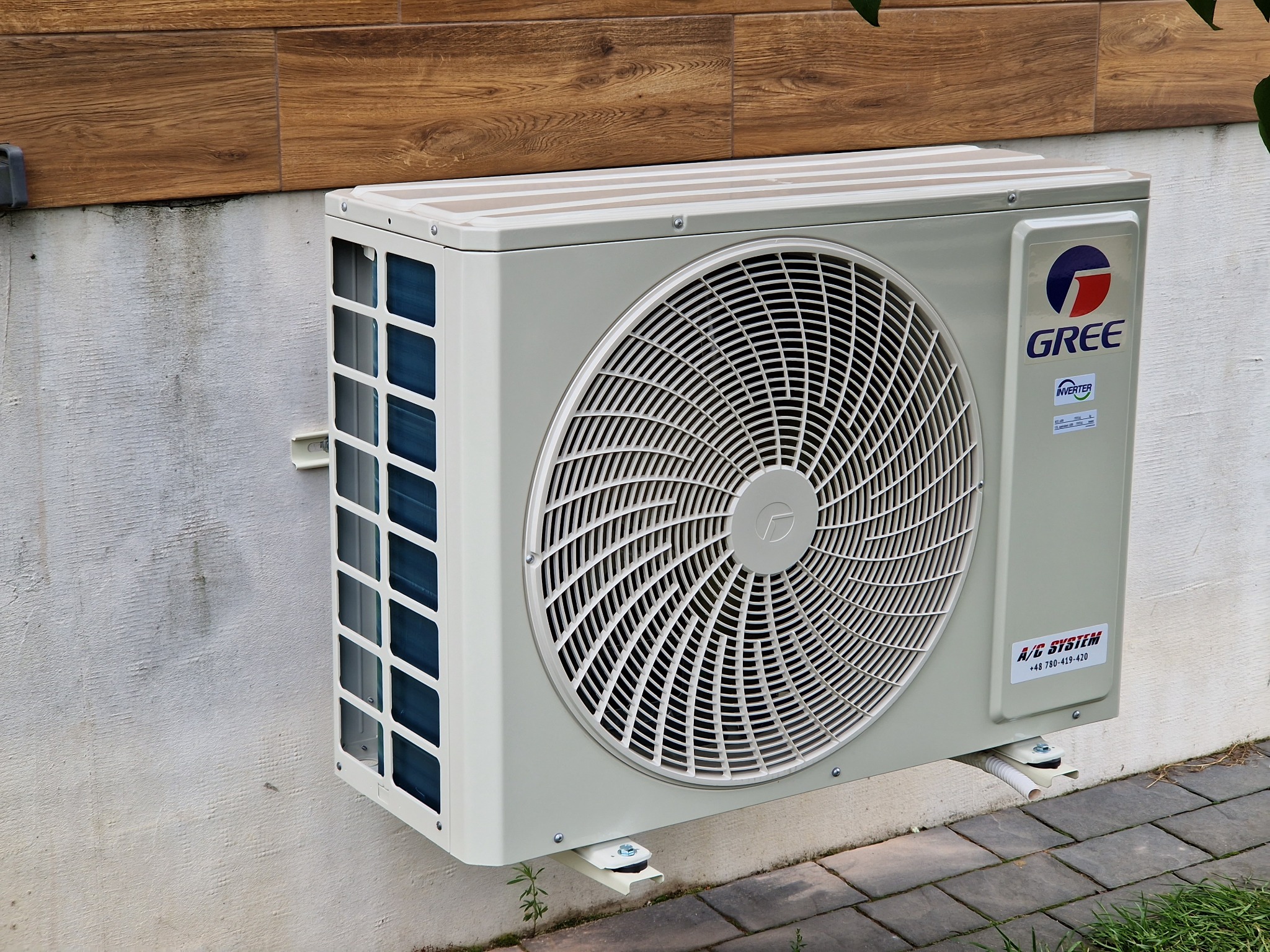 Zewnętrzna jednostka klimatyzacji marki Gree, zamontowana na jasnej ścianie budynku z drewnianą okładziną, widoczne mocowania i fragment chodnika z kostki brukowej.