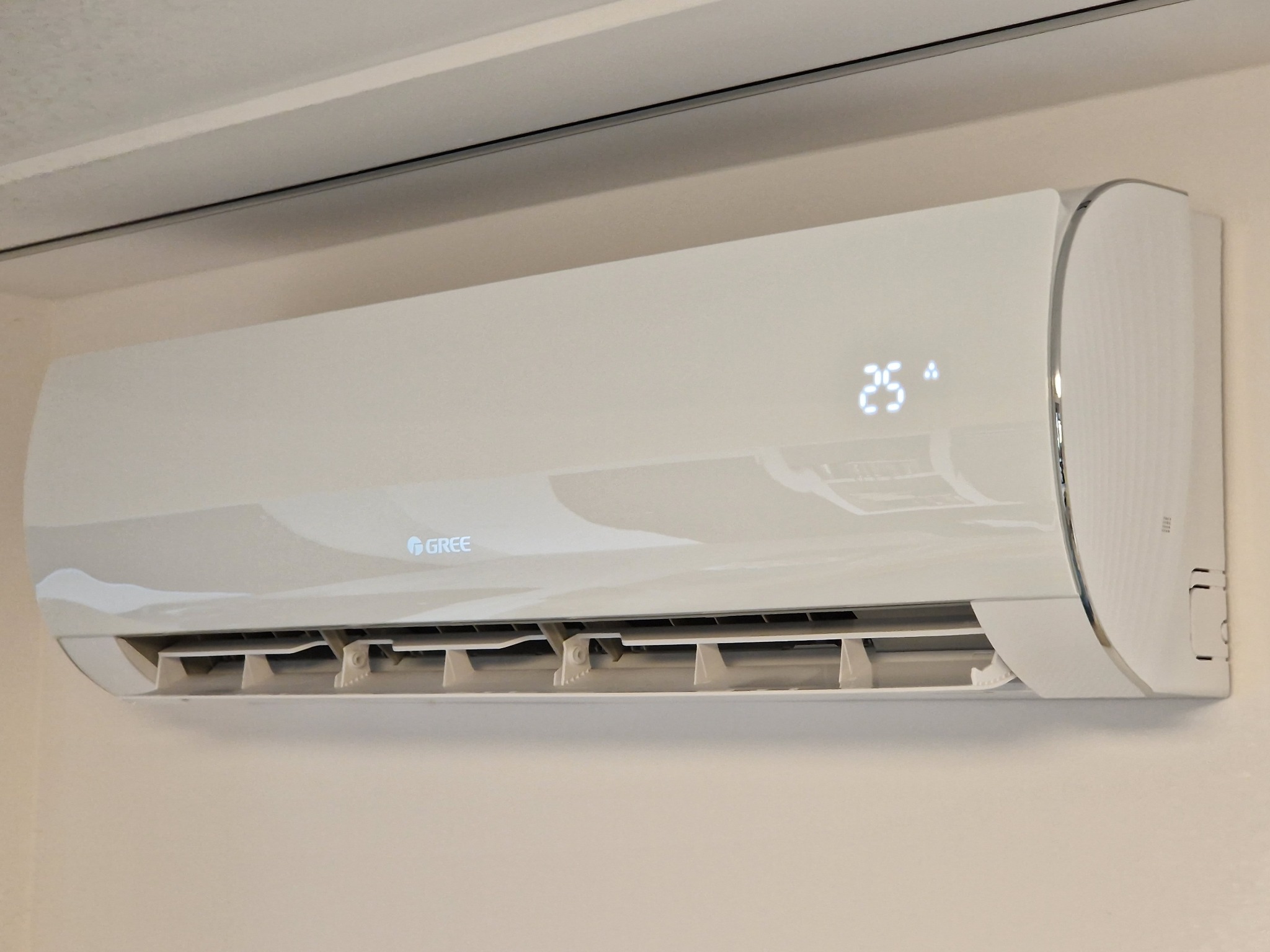 Biały klimatyzator marki Gree zamontowany na ścianie, wyświetlający temperaturę 25 stopni Celsjusza, z widocznymi żaluzjami wylotu powietrza.