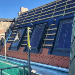 Montaż okien dachowych w trakcie budowy dachu z widocznymi łatami i membraną, część dachu pokryta czerwoną dachówką karpiówką.