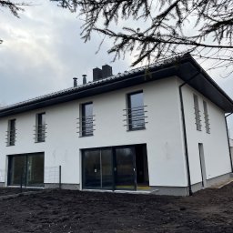 MF Pro Deweloper Michał Fijałka - Nowoczesny, dwukondygnacyjny dom z białymi ścianami i czarnym dachem, z widocznymi oknami z metalowymi balustradami i dużymi przesuwnymi drzwiami tarasowymi, otoczony świeżo przekopaną ziemią...