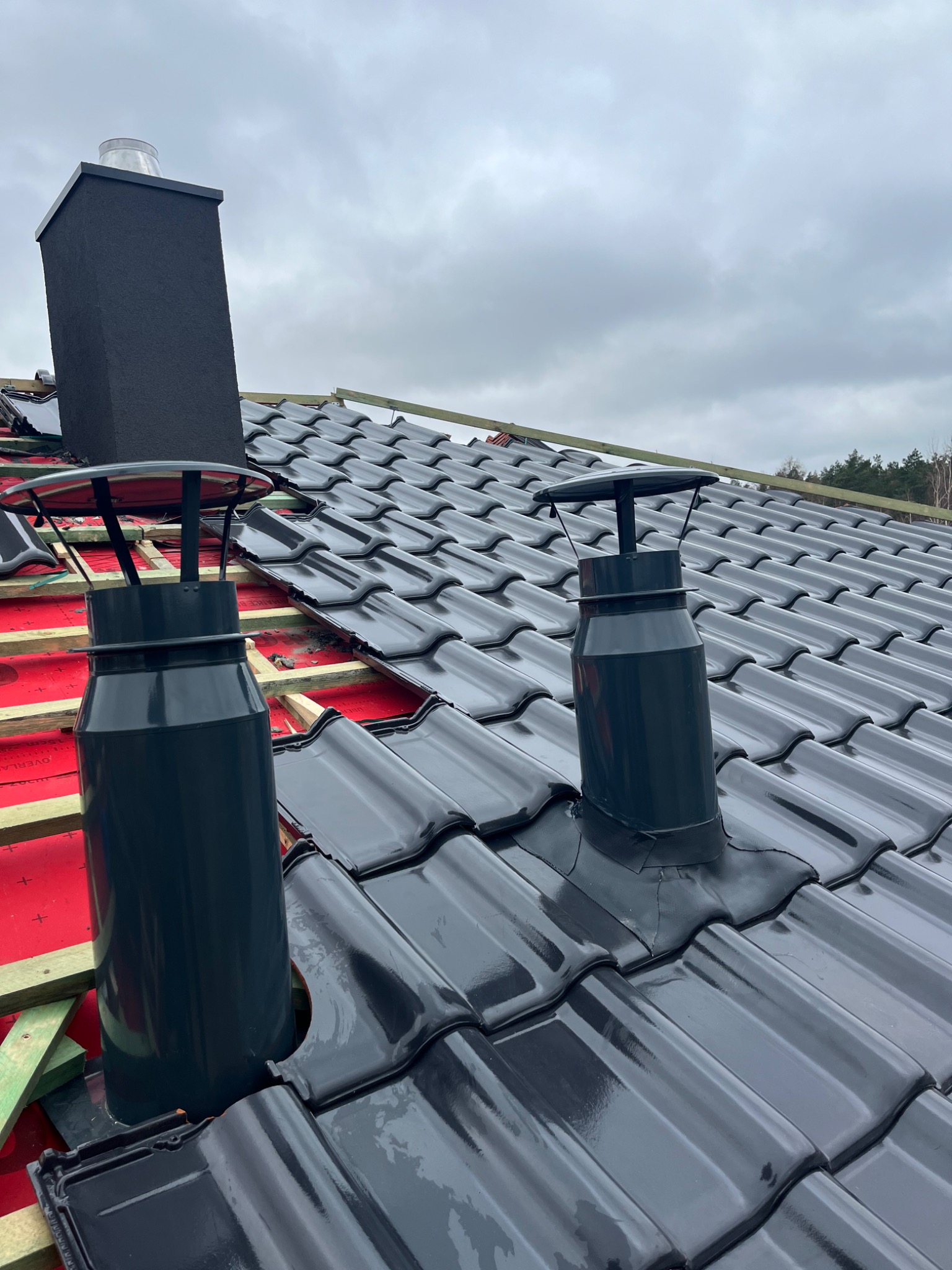 Nowy dach z czarnej dachówki ceramicznej, widoczne kominy wentylacyjne i fragment komina systemowego, montaż na łatach i kontrłatach z czerwoną membraną dachową, pochmurne niebo w tle.