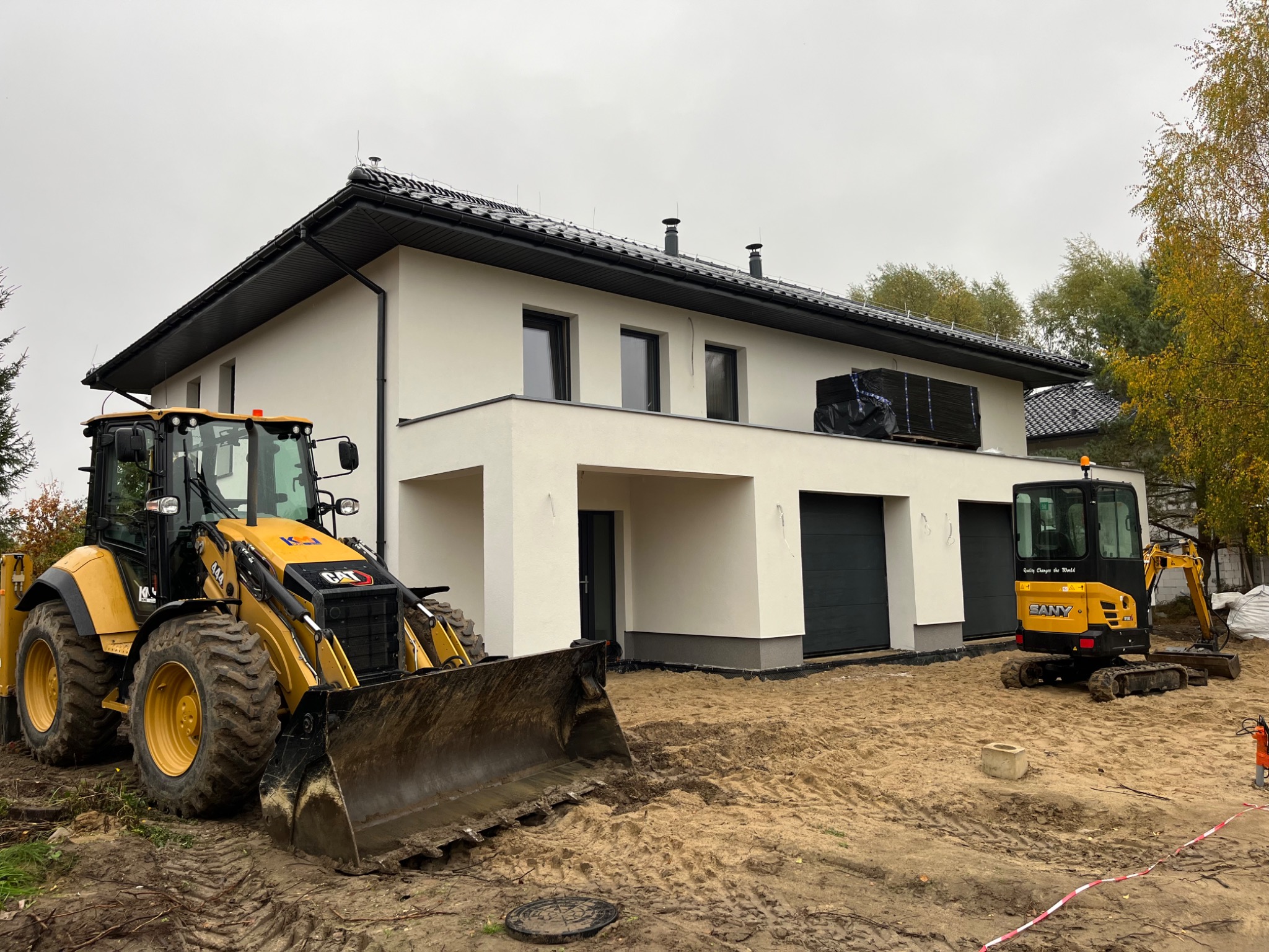 Nowoczesny dom jednorodzinny z ciemną dachówką i dwoma żółtymi maszynami budowlanymi: koparko-ładowarką CAT oraz mini koparką SANY, teren przygotowywany pod przyszłe prace brukarskie.