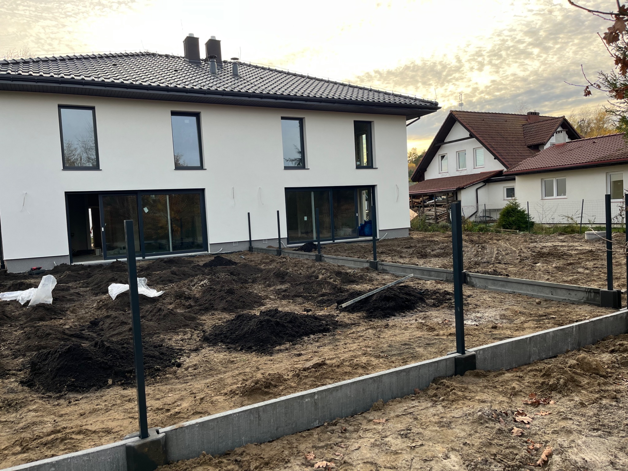 Nowoczesny dom z ciemną dachówką w trakcie budowy, widoczne fundamenty pod kostkę brukową i przygotowane podłoże z czarną ziemią.