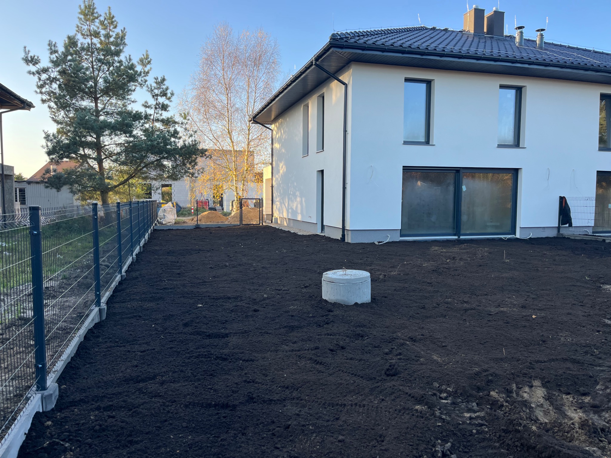 Przygotowany teren pod trawnik z widocznym włazem kanalizacyjnym, ogrodzeniem panelowym i fragmentem elewacji nowego domu z ciemną dachówką.