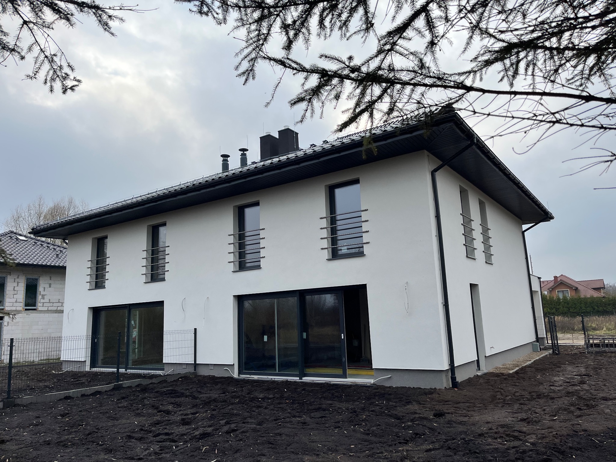 Nowoczesny, dwukondygnacyjny dom z białymi ścianami i czarnym dachem, z widocznymi oknami z metalowymi balustradami i dużymi przesuwnymi drzwiami tarasowymi, otoczony świeżo przekopaną ziemią...