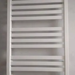 Biały, drabinkowy grzejnik łazienkowy zamontowany na szarej ścianie, z widocznymi zaworami przyłączeniowymi.