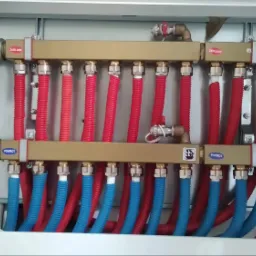 Rozdzielacz ogrzewania podłogowego z rurami w kolorze czerwonym i niebieskim, zamontowany w białej szafce, widoczne zawory z napisem 'Trinity'.