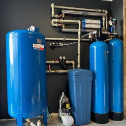 Drill Solutions Sp. Zo.o. - Kompletna instalacja uzdatniania wody w pomieszczeniu z ciemnoszarą ścianą, obejmująca zbiornik przeponowy WIMEST, filtry kolumnowe, lampy UV i mały kompresor.