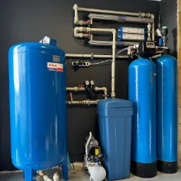 Kompletna instalacja uzdatniania wody w pomieszczeniu z ciemnoszarą ścianą, obejmująca zbiornik przeponowy WIMEST, filtry kolumnowe, lampy UV i mały kompresor.