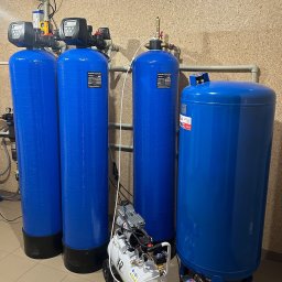 Drill Solutions Sp. Zo.o. - Instalacja stacji uzdatniania wody z czterema niebieskimi zbiornikami ciśnieniowymi i białym kompresorem obok, w pomieszczeniu z wykończeniem z płyty OSB.