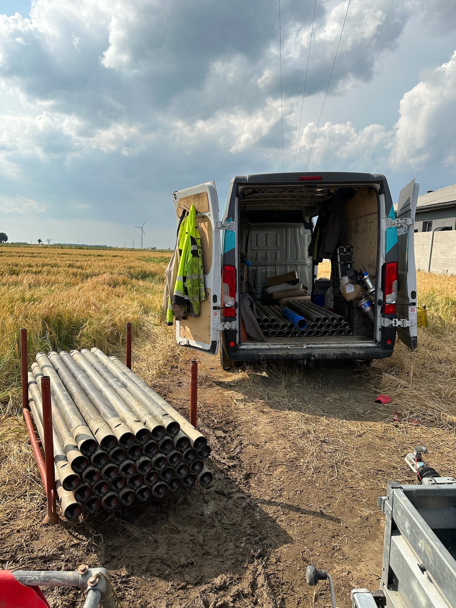 Stos rur w metalowej ramie obok otwartego furgonetki z widocznym wyposażeniem i narzędziami na tle pola i wiatraka w oddali.