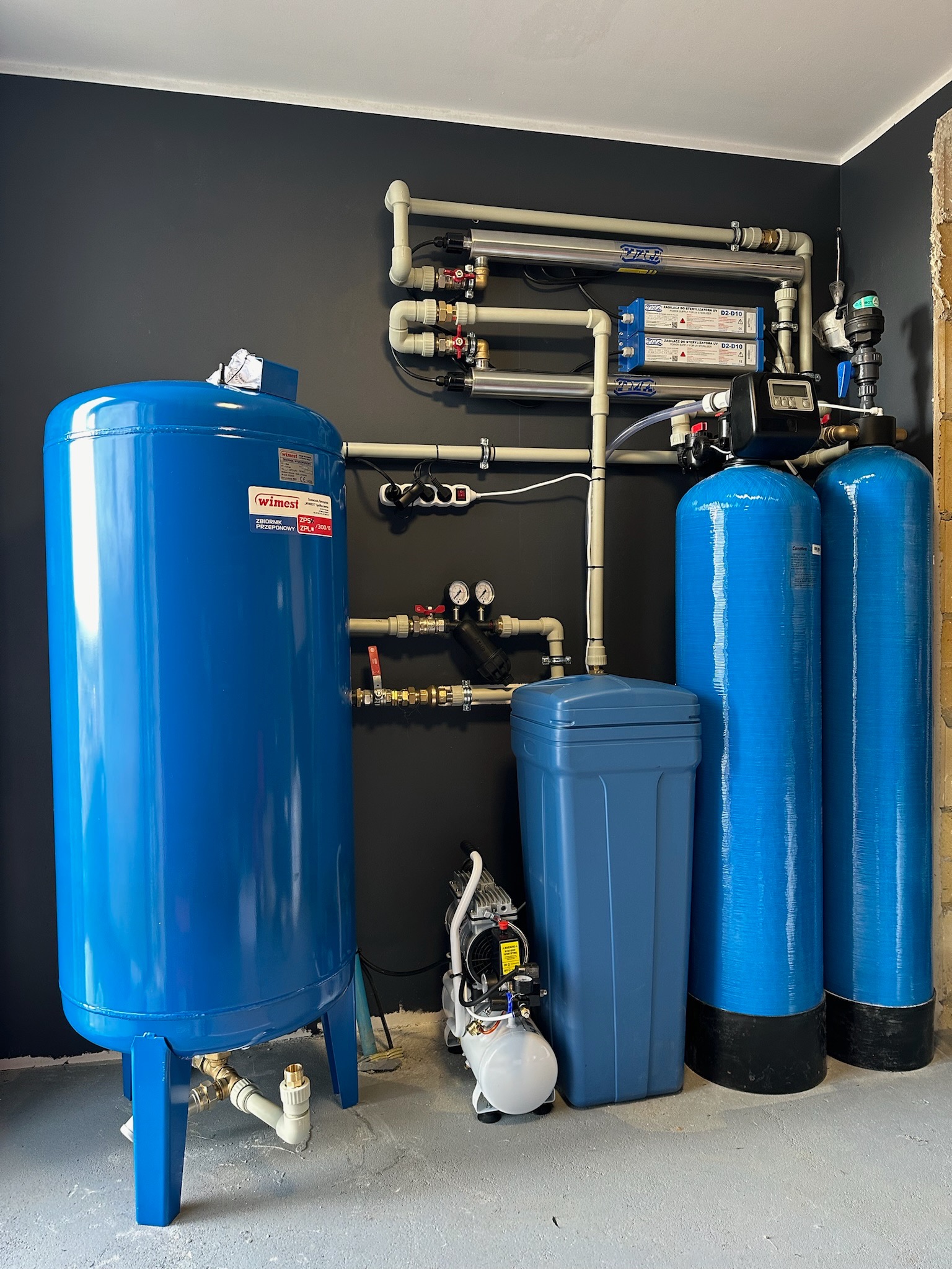 Kompletna instalacja uzdatniania wody w pomieszczeniu z ciemnoszarą ścianą, obejmująca zbiornik przeponowy WIMEST, filtry kolumnowe, lampy UV i mały kompresor.