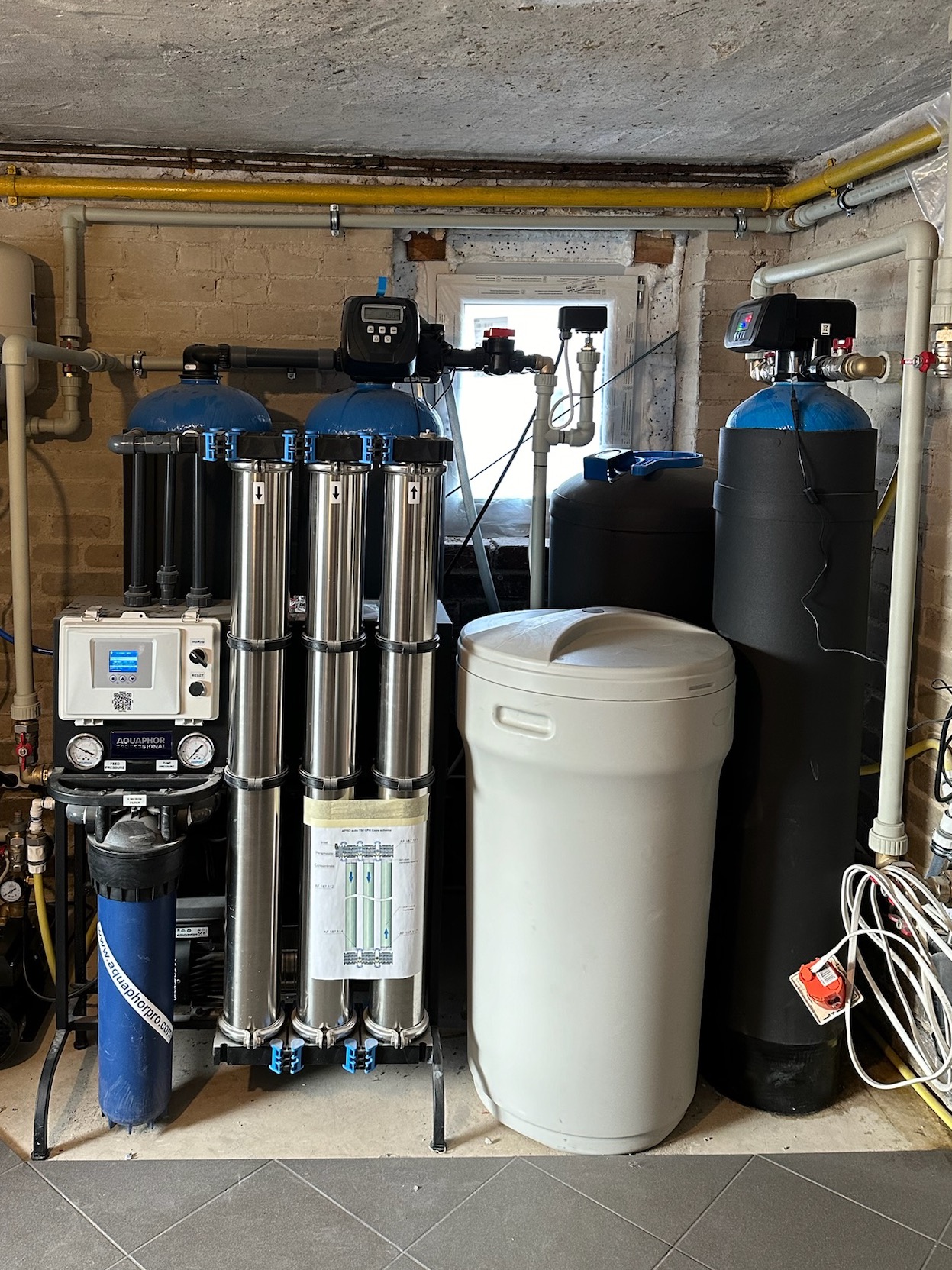 Zestaw urządzeń do filtracji wody z kilkoma cylindrycznymi filtrami ze stali nierdzewnej, zbiornikiem z tworzywa sztucznego i elektronicznym sterownikiem, zainstalowany w pomieszczeniu z ceglanymi...