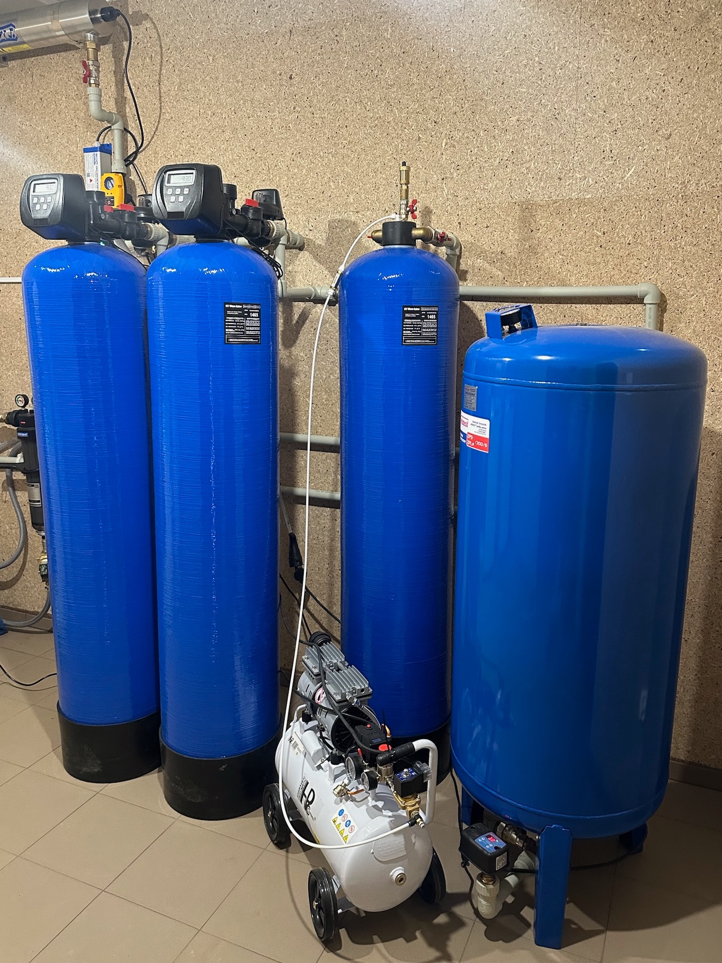 Instalacja stacji uzdatniania wody z czterema niebieskimi zbiornikami ciśnieniowymi i białym kompresorem obok, w pomieszczeniu z wykończeniem z płyty OSB.