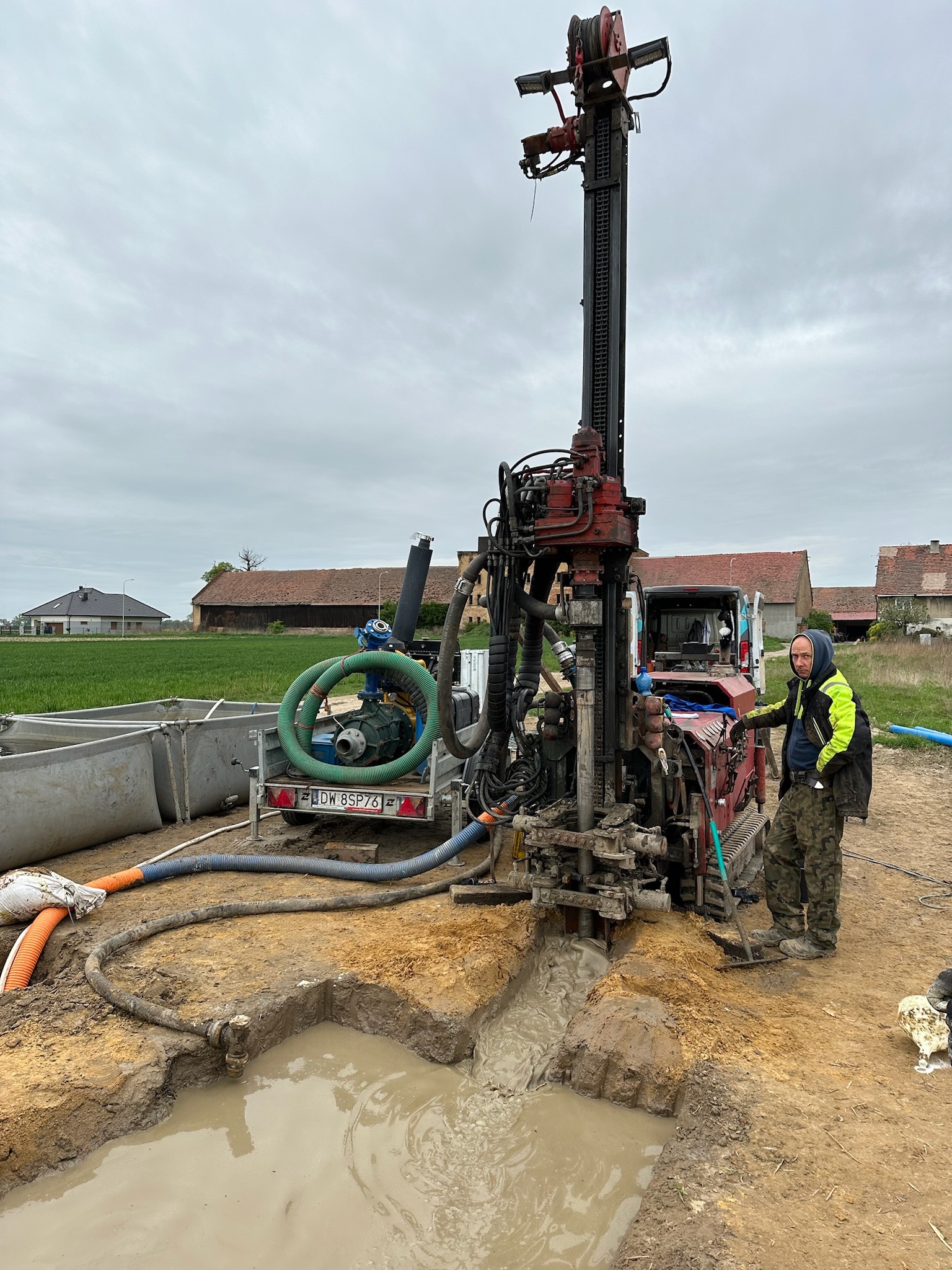 Wiertnica do studni głębinowej w trakcie pracy, z widocznym osprzętem i pracownikiem w odblaskowej kurtce, teren błotnisty, w tle zabudowania gospodarcze.