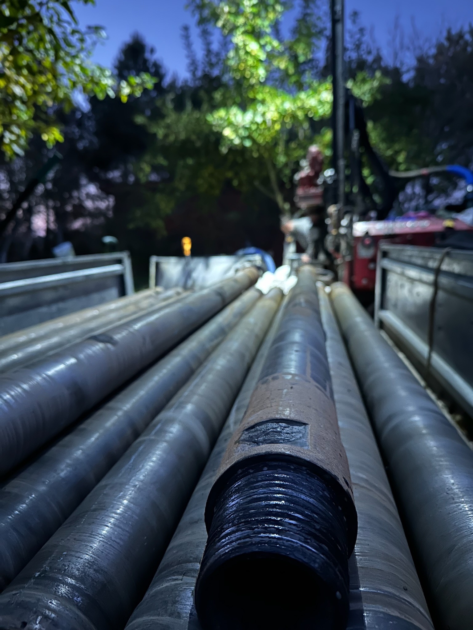 Rury wiertnicze z gwintem na pierwszym planie, maszyna wiertnicza i operator w tle, oświetlone lampami w nocy.