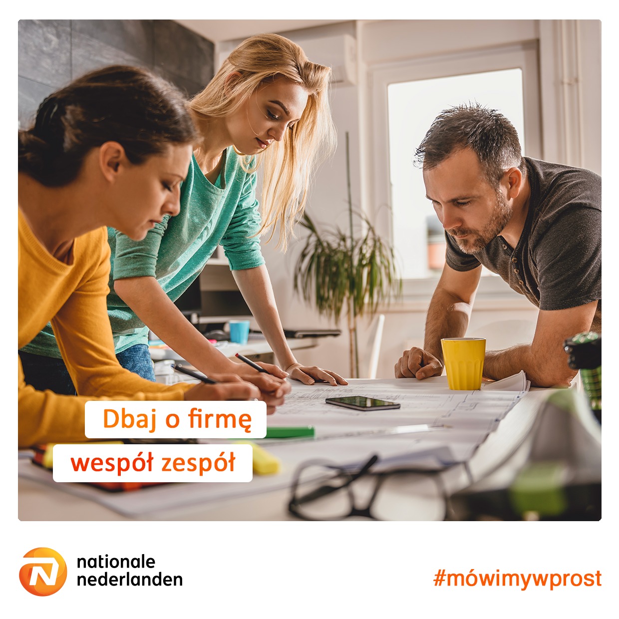 Trzy osoby pochylone nad stołem analizują dokumenty, na stole leżą okulary, telefon i żółty kubek, w tle widoczne biuro.