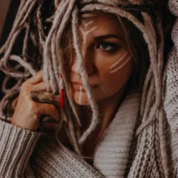 Portret kobiety z dredami, z delikatnym makijażem i białymi geometrycznymi wzorami na twarzy, ubrana w gruby sweter z dzianiny.