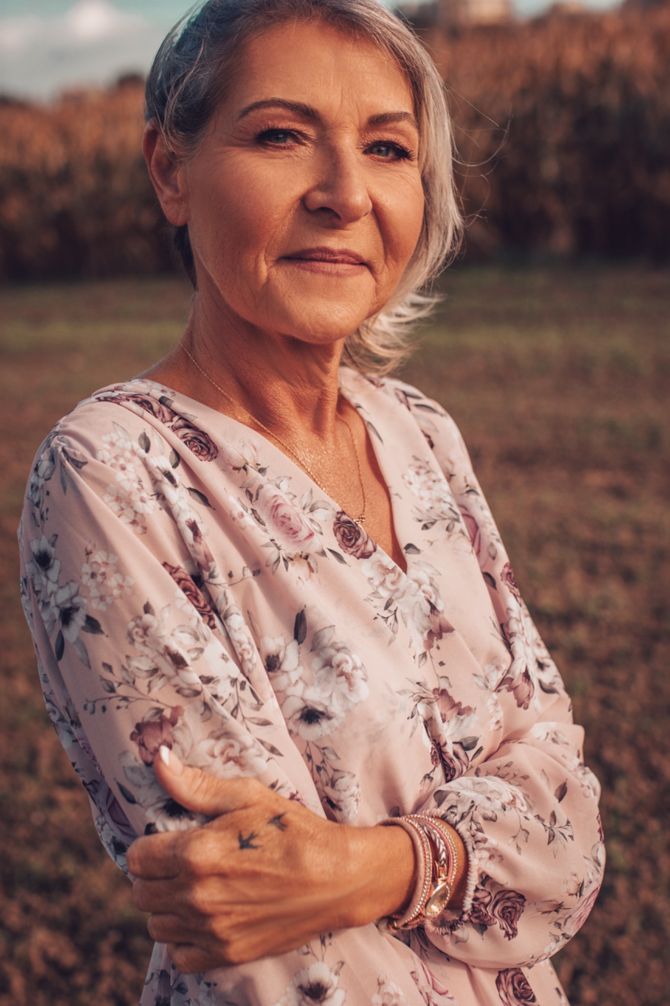 Portret dojrzałej kobiety z siwymi włosami w kwiecistej sukience, z delikatną biżuterią i tatuażem dwóch ptaków na dłoni, na tle rozmytej zieleni.