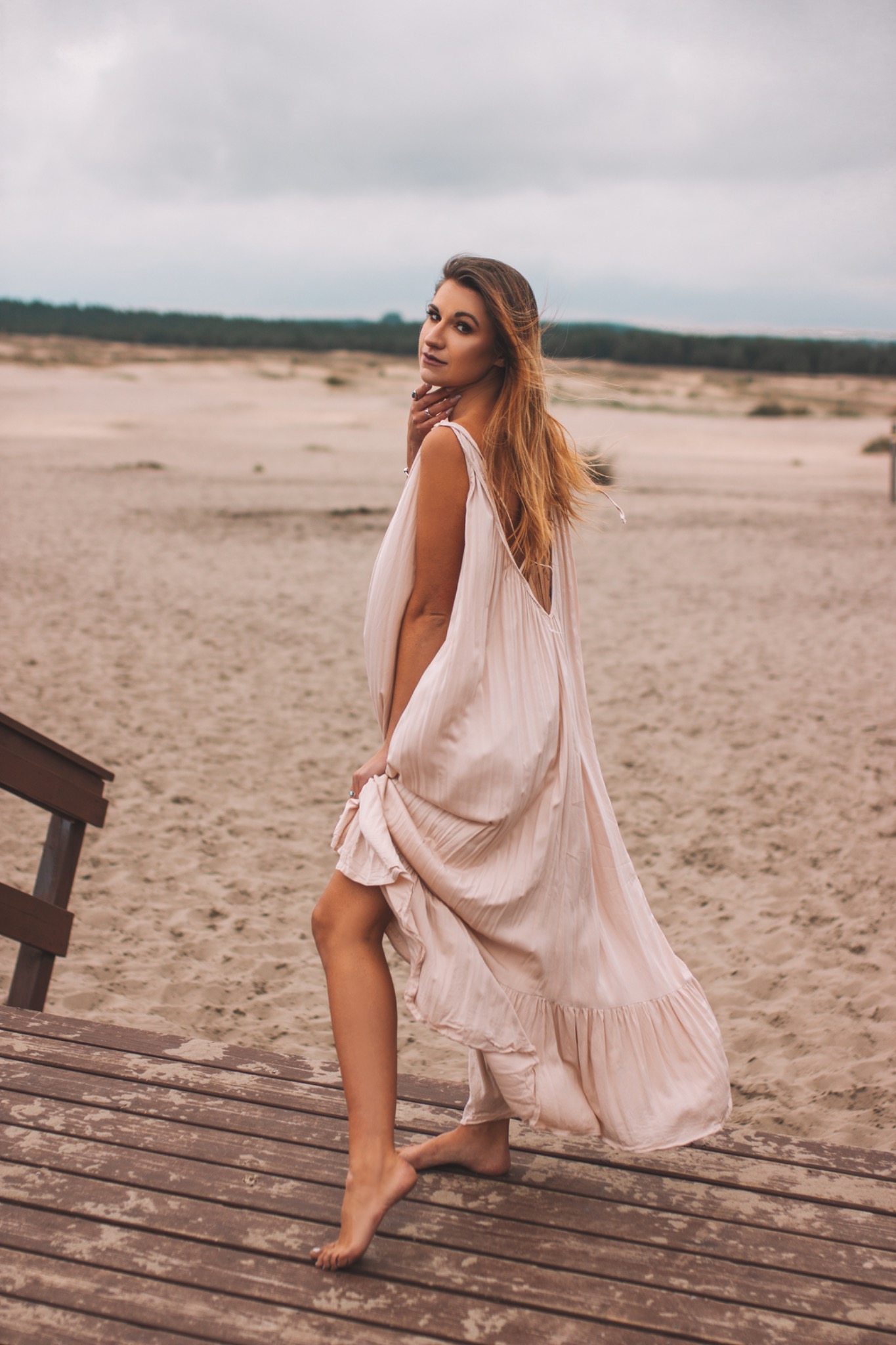 Kobieta w zwiewnej, jasnoróżowej sukience, stojąca na drewnianym podeście na plaży, spogląda przez ramię na fotografa.