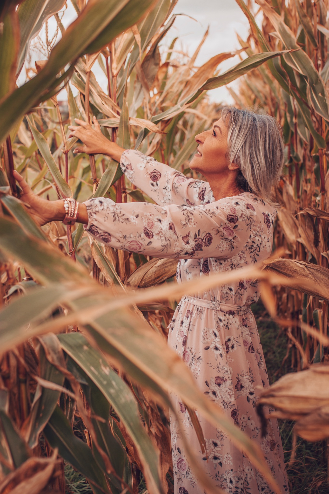 Kobieta w kwiecistej sukience, z uśmiechem na twarzy, pozuje w polu kukurydzy, otoczona wysokimi łodygami i liśćmi.