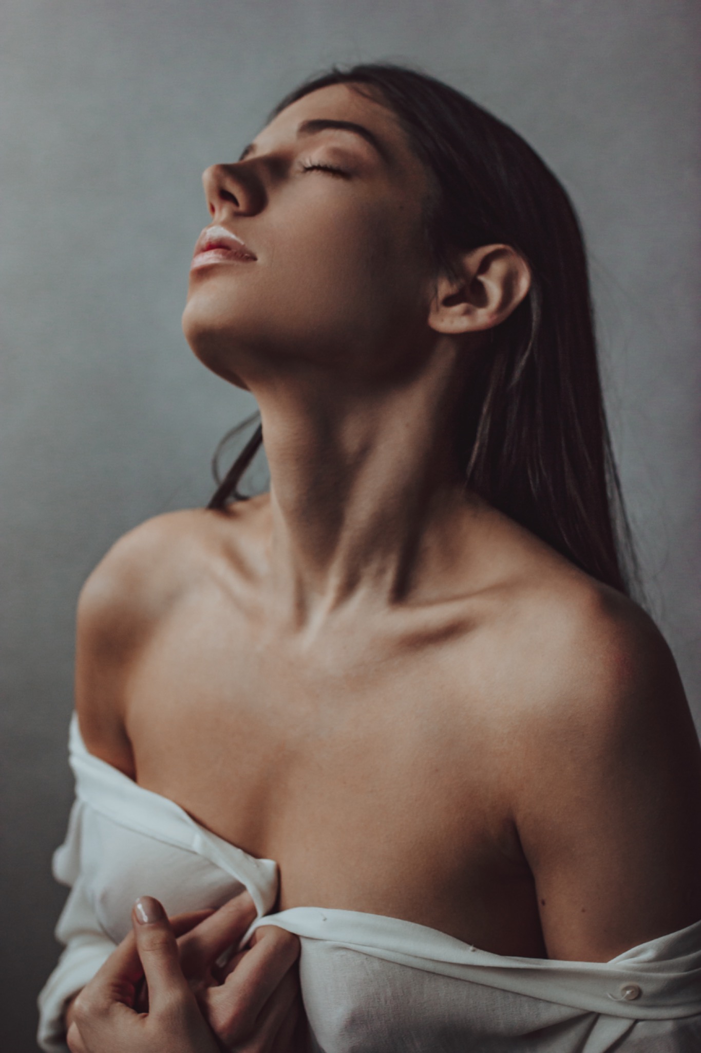 Portret kobiety z zamkniętymi oczami, rozpięta biała koszula, delikatne światło padające na twarz i szyję, neutralne tło.