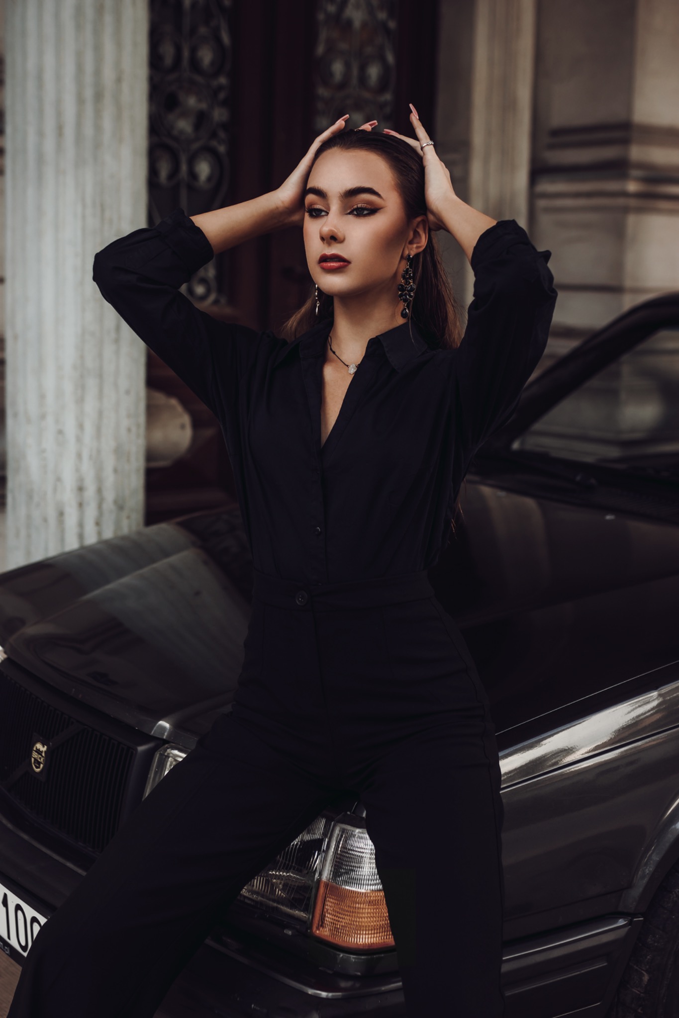 Kobieta w czarnym kombinezonie pozuje oparta o maskę ciemnego samochodu, z rękami uniesionymi nad głową i kolumnami w tle.