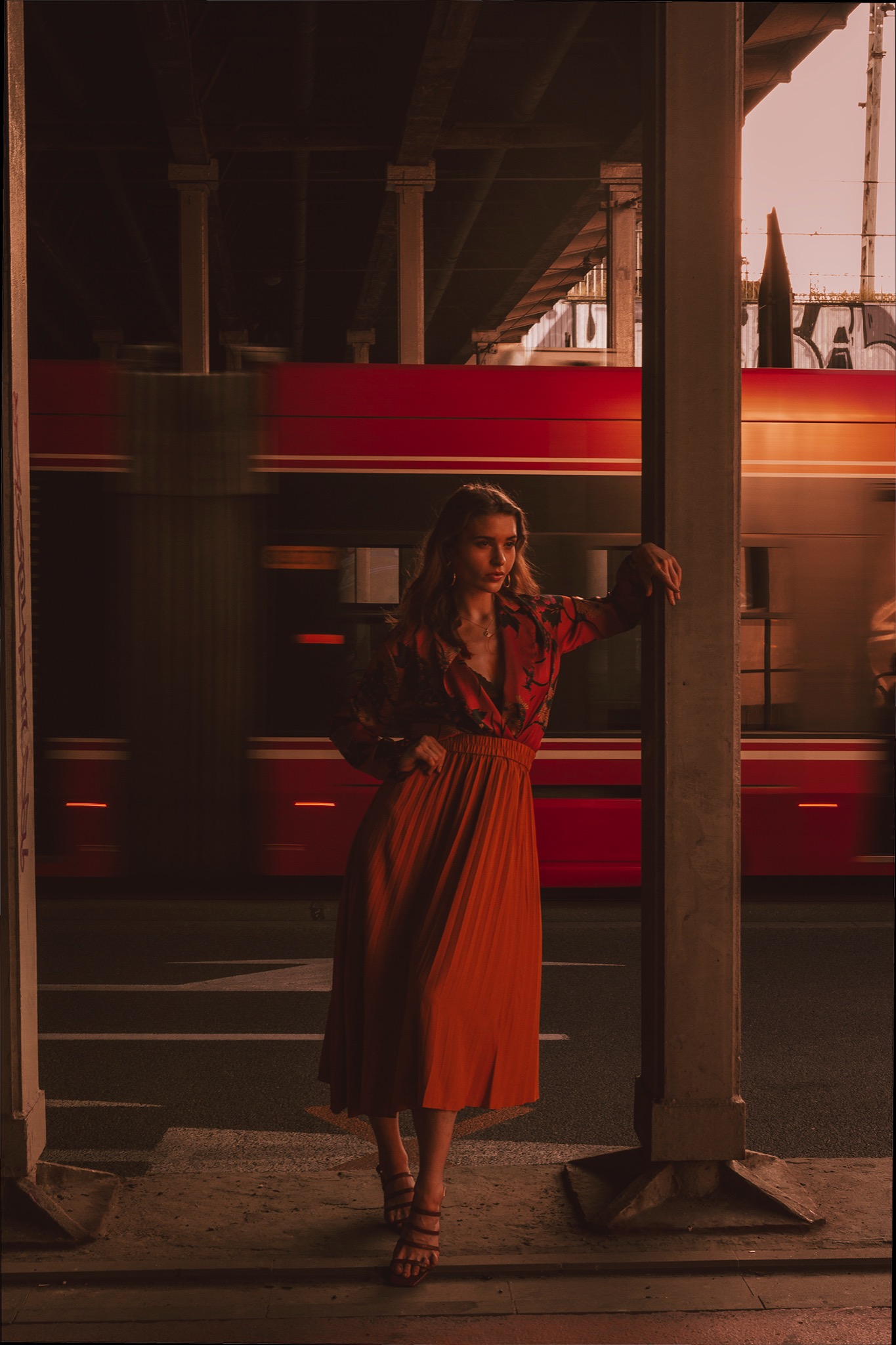 Młoda kobieta w kwiecistej bluzce i plisowanej spódnicy w kolorze rdzy, oparta o filar na tle rozmazanego, czerwonego tramwaju, ujęcie w miejskiej scenerii z ciepłym oświetleniem.
