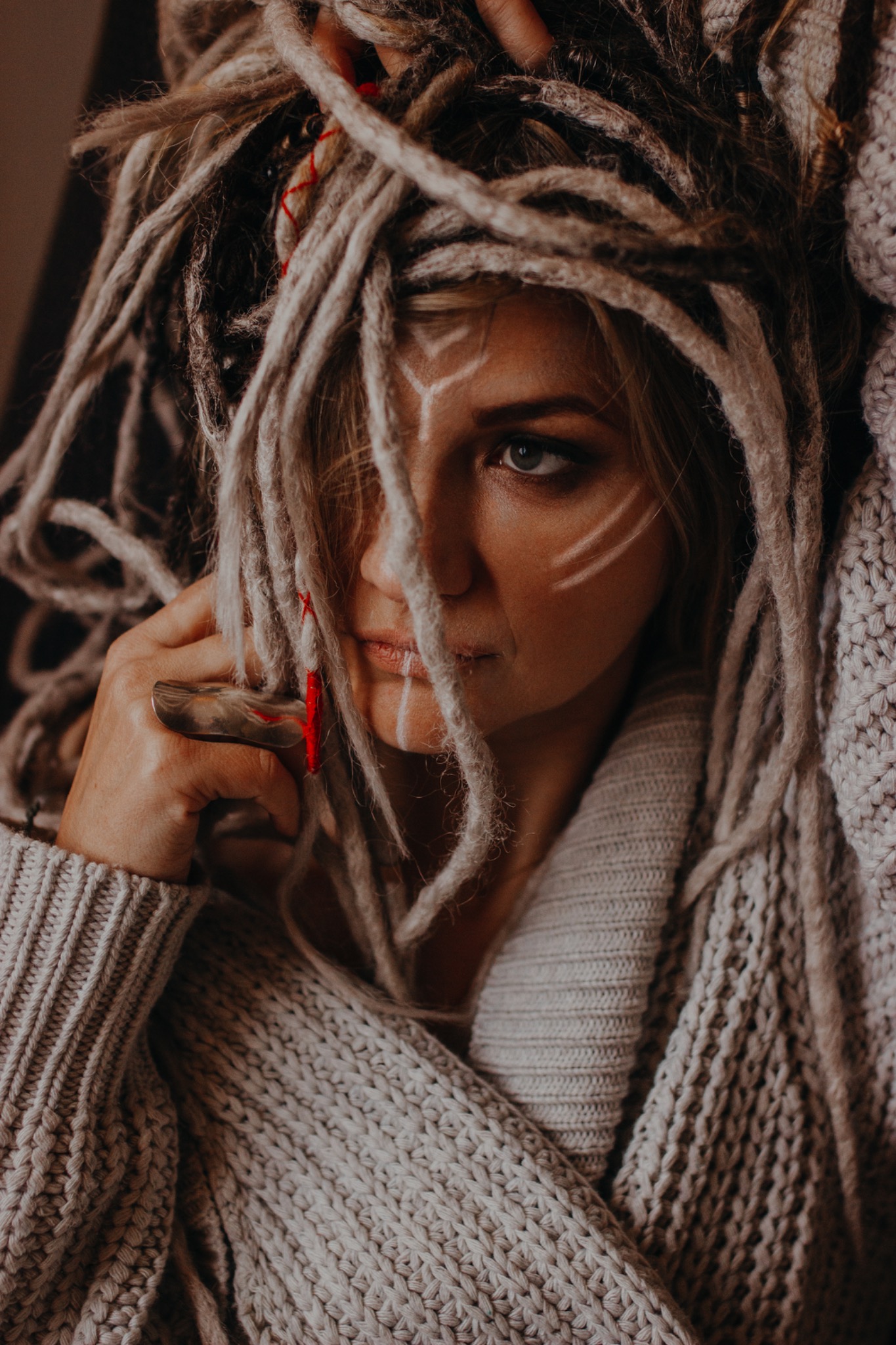 Portret kobiety z dredami, z delikatnym makijażem i białymi geometrycznymi wzorami na twarzy, ubrana w gruby sweter z dzianiny.
