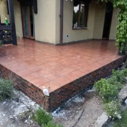 Wykończony taras z brązowych płytek ceramicznych na podwyższeniu z cegieł, widoczne zielone krzewy i fragment budynku z oknami.