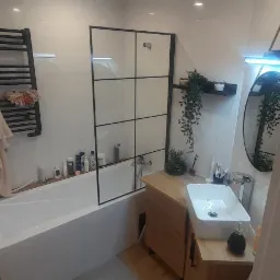 Nowoczesna łazienka z białą armaturą, kabiną prysznicową typu walk-in z czarnymi profilami, drewnianą szafką pod umywalką nablatową i dekoracyjnymi roślinami doniczkowymi.