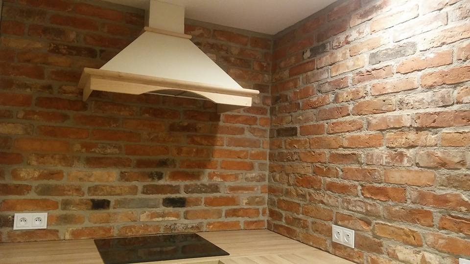 Kuchnia z ceglaną ścianą, drewnianym blatem i okapem w stylu retro, gniazdka elektryczne zamontowane na cegle.