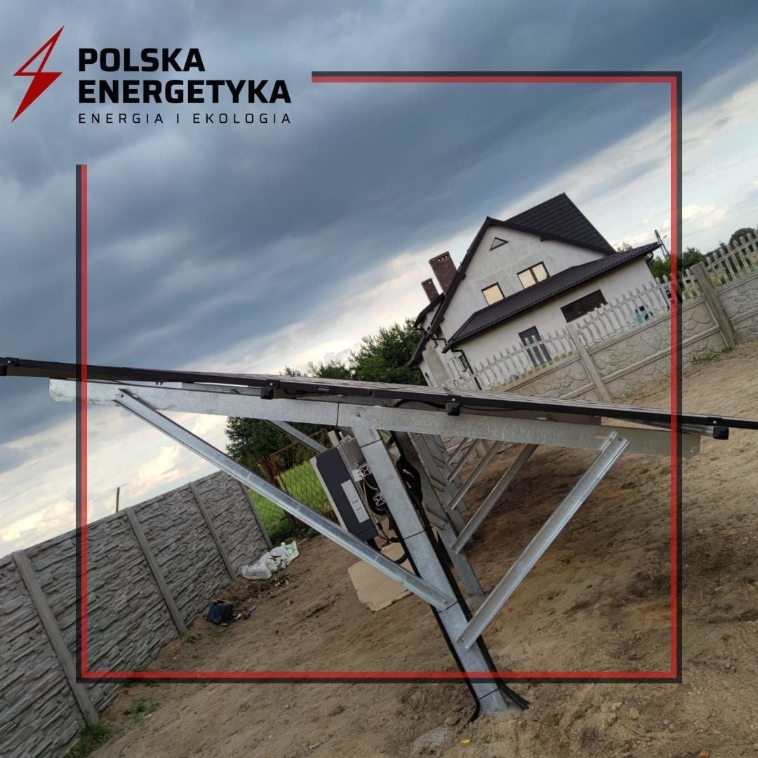 Konstrukcja wsporcza dla paneli słonecznych na tle domu jednorodzinnego i pochmurnego nieba, widoczne elementy montażowe i okablowanie.