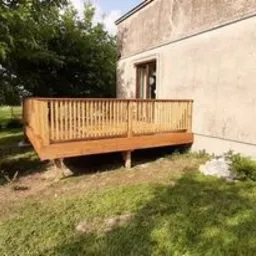 Nowo wybudowany taras drewniany przylegający do ściany budynku z widoczną roślinnością w tle, wykonany z jasnego drewna i zabezpieczony lakierem. Taras umiejscowiony na lekko pochyłym terenie.