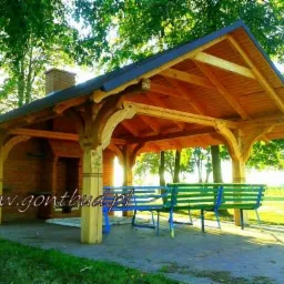 Drewniana altana z ceglanym kominkiem, oświetlona popołudniowym słońcem, z dwoma ławkami w kolorach niebieskim i zielonym.