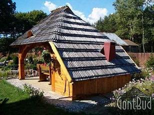 Altana ogrodowa kryta gontem drewnianym, z widocznym fragmentem ławki i otoczeniem kwiatów oraz drzew