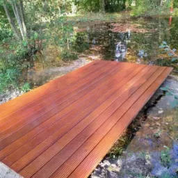 Pomost z desek kompozytowych nad stawem w ogrodzie, widok z niskiej perspektywy, otoczony zielenią.