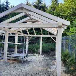 Przemysław Kusowski - Konstrukcja drewnianej altany w trakcie budowy, widoczne rusztowanie i elementy mocowania w ziemi, tło z zielenią.